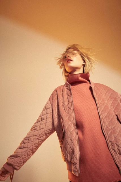 Blond woman with windblown hair poses in a pink quilted jacket and turtleneck against a peach background in a fashion portrait.