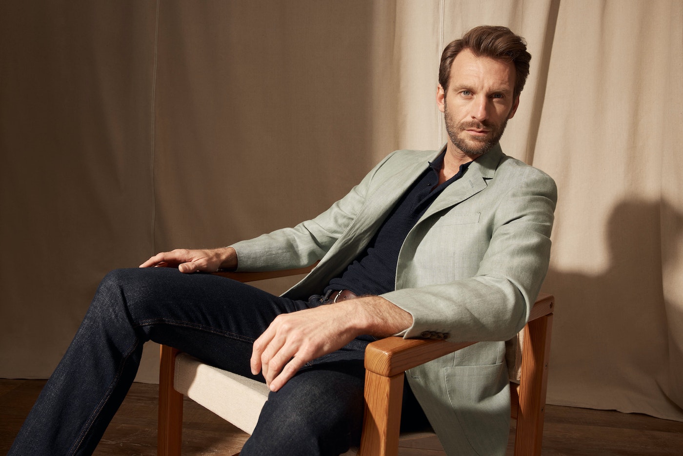 Portrait of an adult male seated in a chair wearing a light green jacket, dark blue shirt, and dark jeans in a neutral indoor setting.