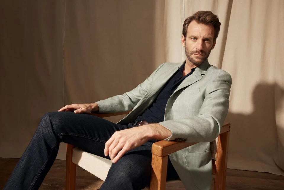 Portrait of an adult male seated in a chair wearing a light green jacket, dark blue shirt, and dark jeans in a neutral indoor setting.