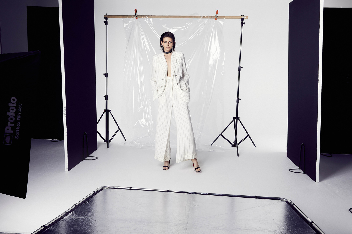 Elegant adult female wearing a white pantsuit poses in a photo studio with soft lighting and studio equipment in the background.