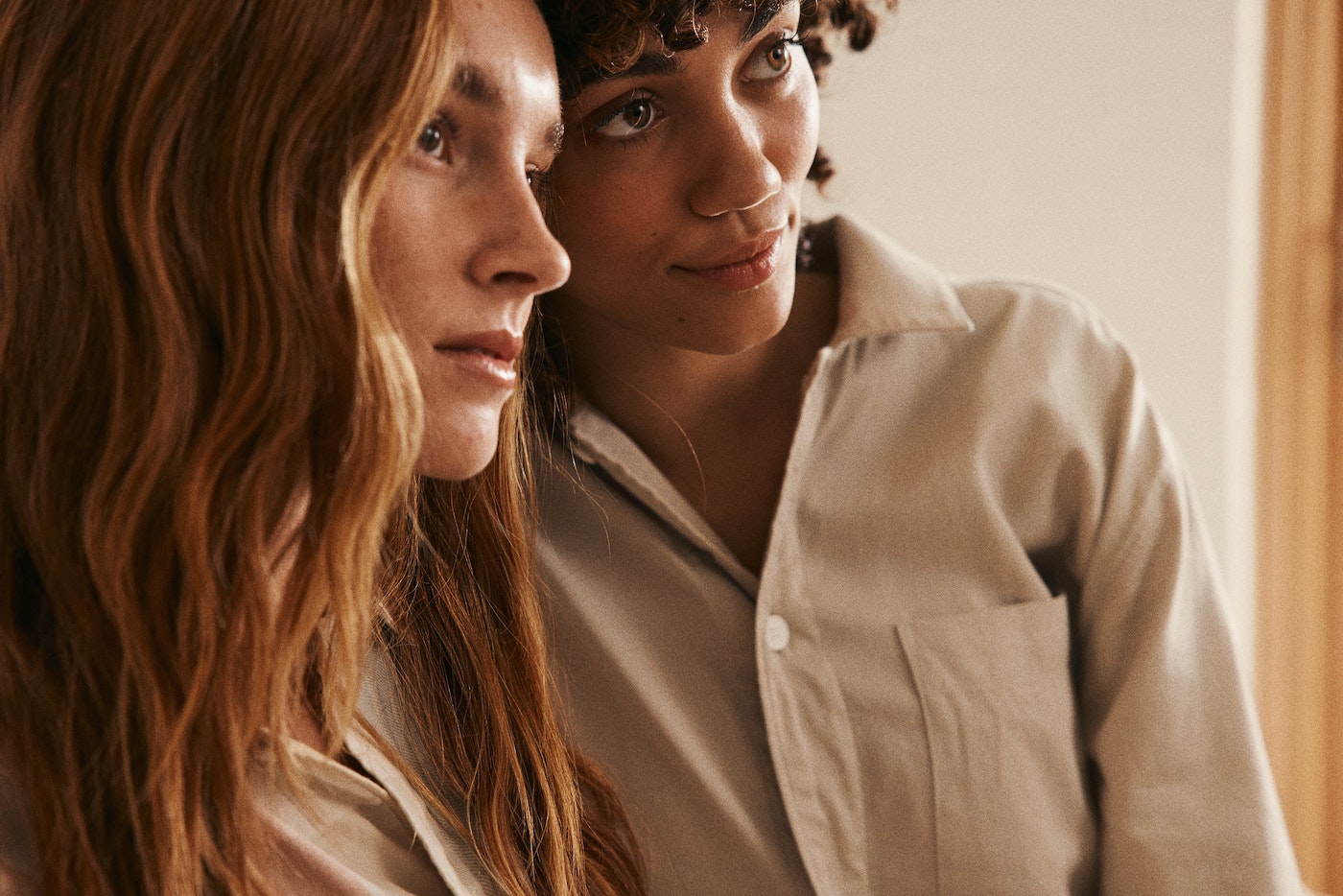 Two young women with auburn and curly brunette hair pose indoors, wearing casual collared shirts in a medium studio shot.