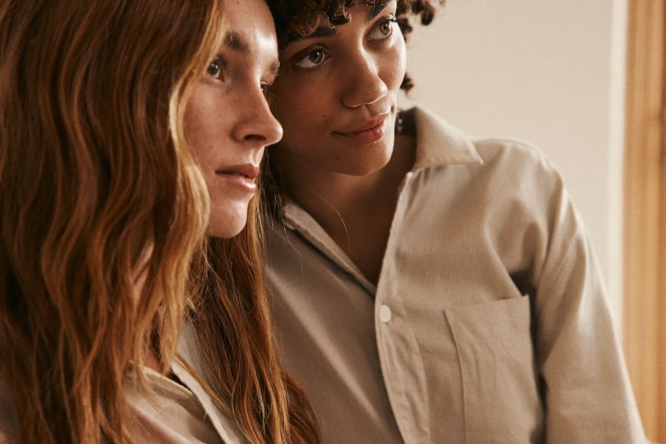 Two young women with auburn and curly brunette hair pose indoors, wearing casual collared shirts in a medium studio shot.
