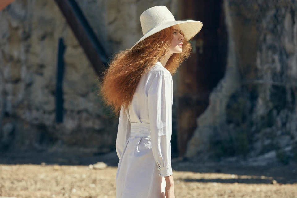 A red-haired woman in a white dress and sunhat gazes to the left, standing outdoors against a textured, earthy-toned backdrop.