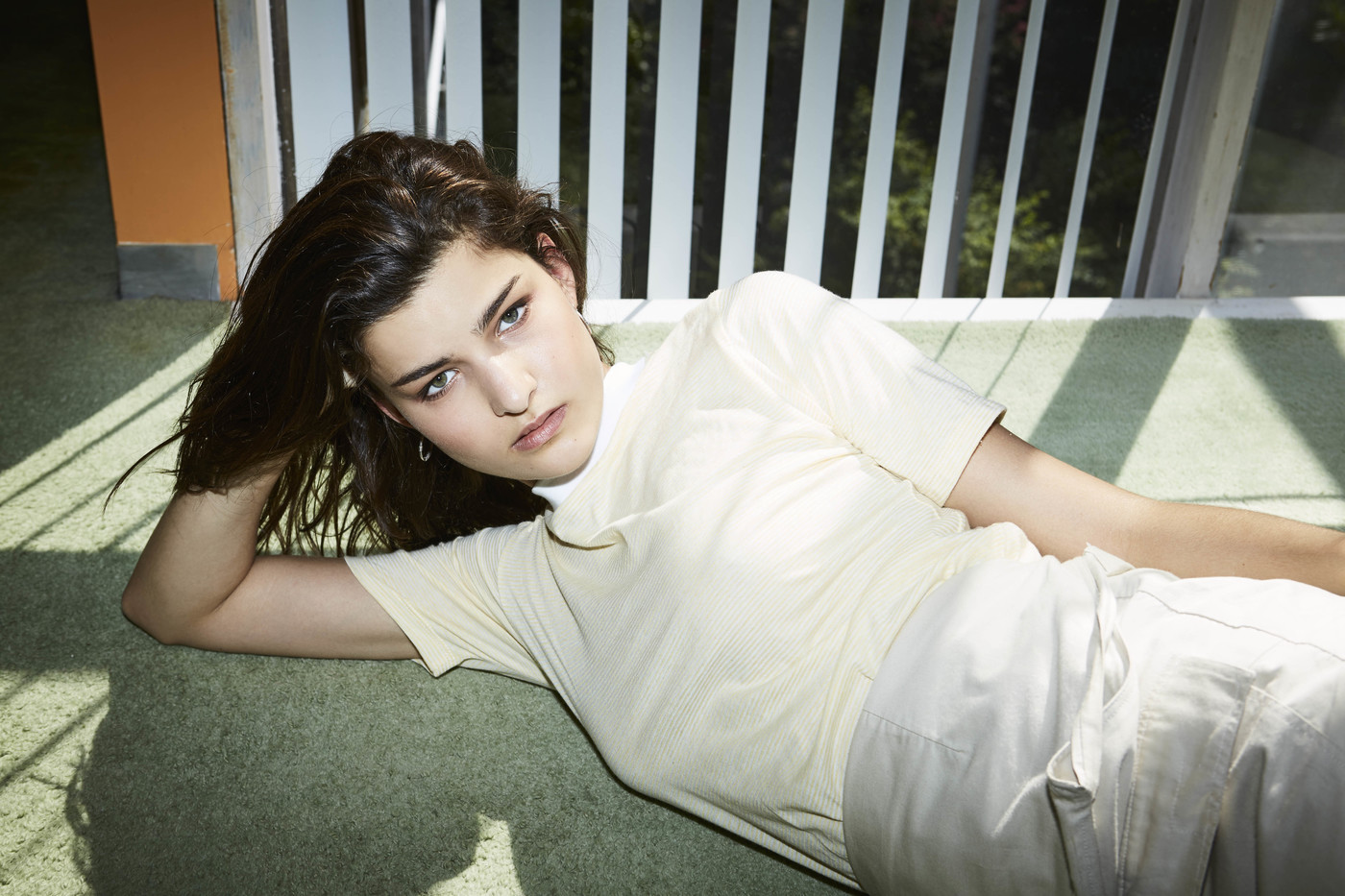 Young woman with dark hair reclining on green carpeted floor near vertical white window blinds by photographer Michelle Tran
