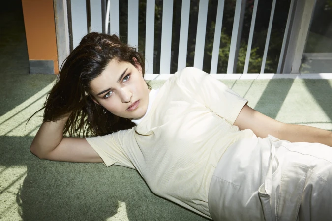 Young woman with dark hair reclining on green carpeted floor near vertical white window blinds by photographer Michelle Tran