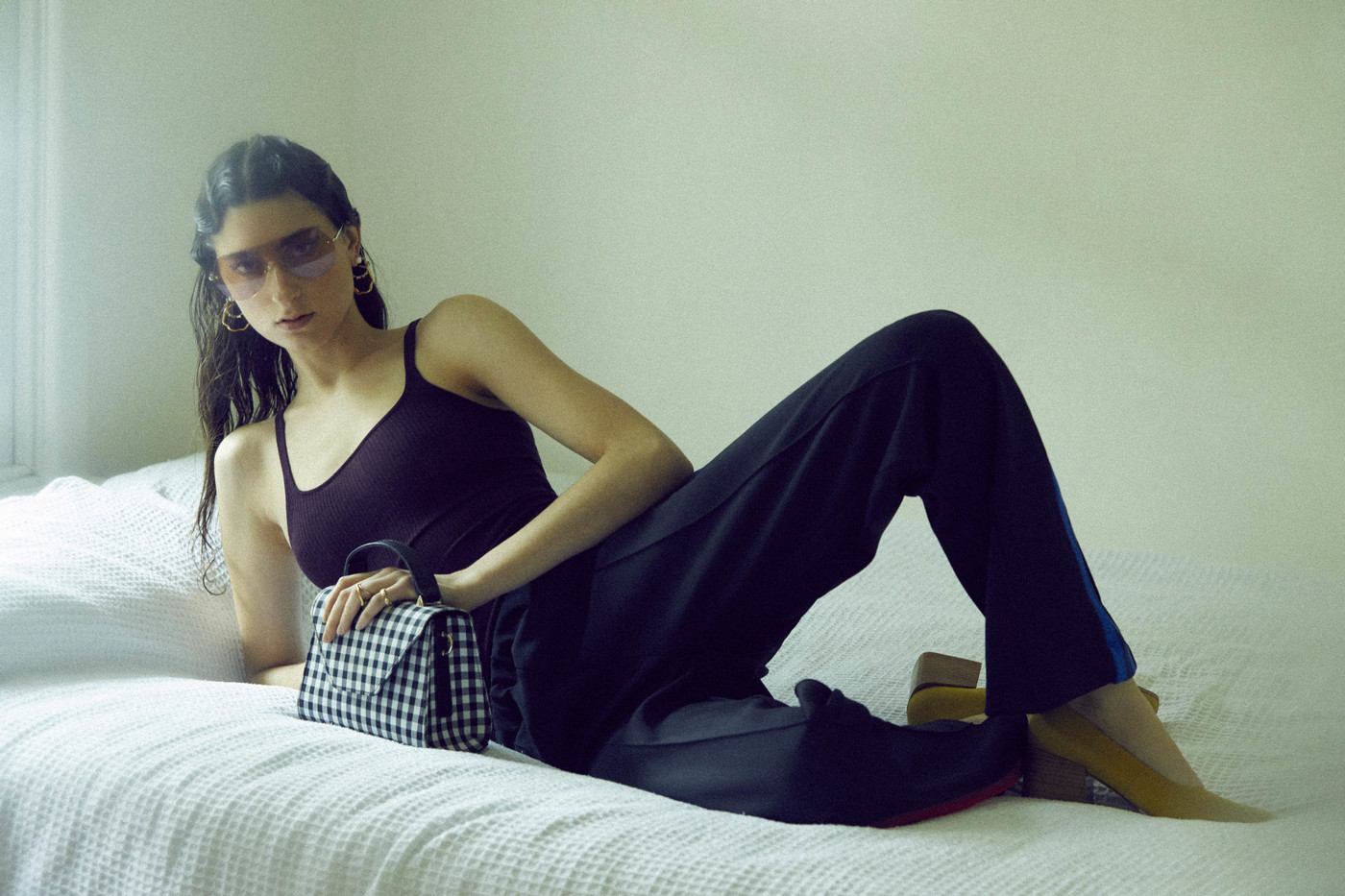 Indoor shot of a woman reclining in black top and pants, holding a black and white gingham handbag, with yellow shoes.