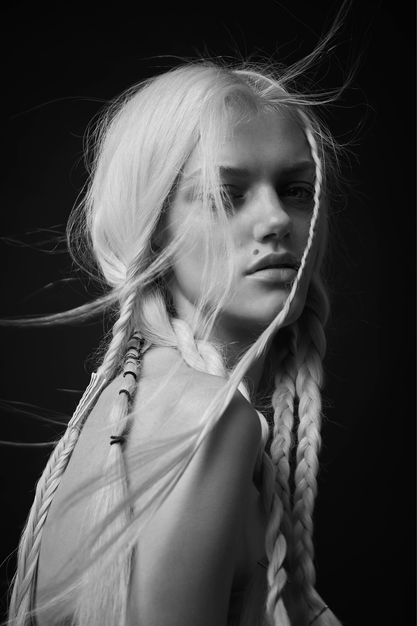 Portrait of a blonde woman with braided hair and loose strands blowing in the wind against a dark background by Michelle Tran