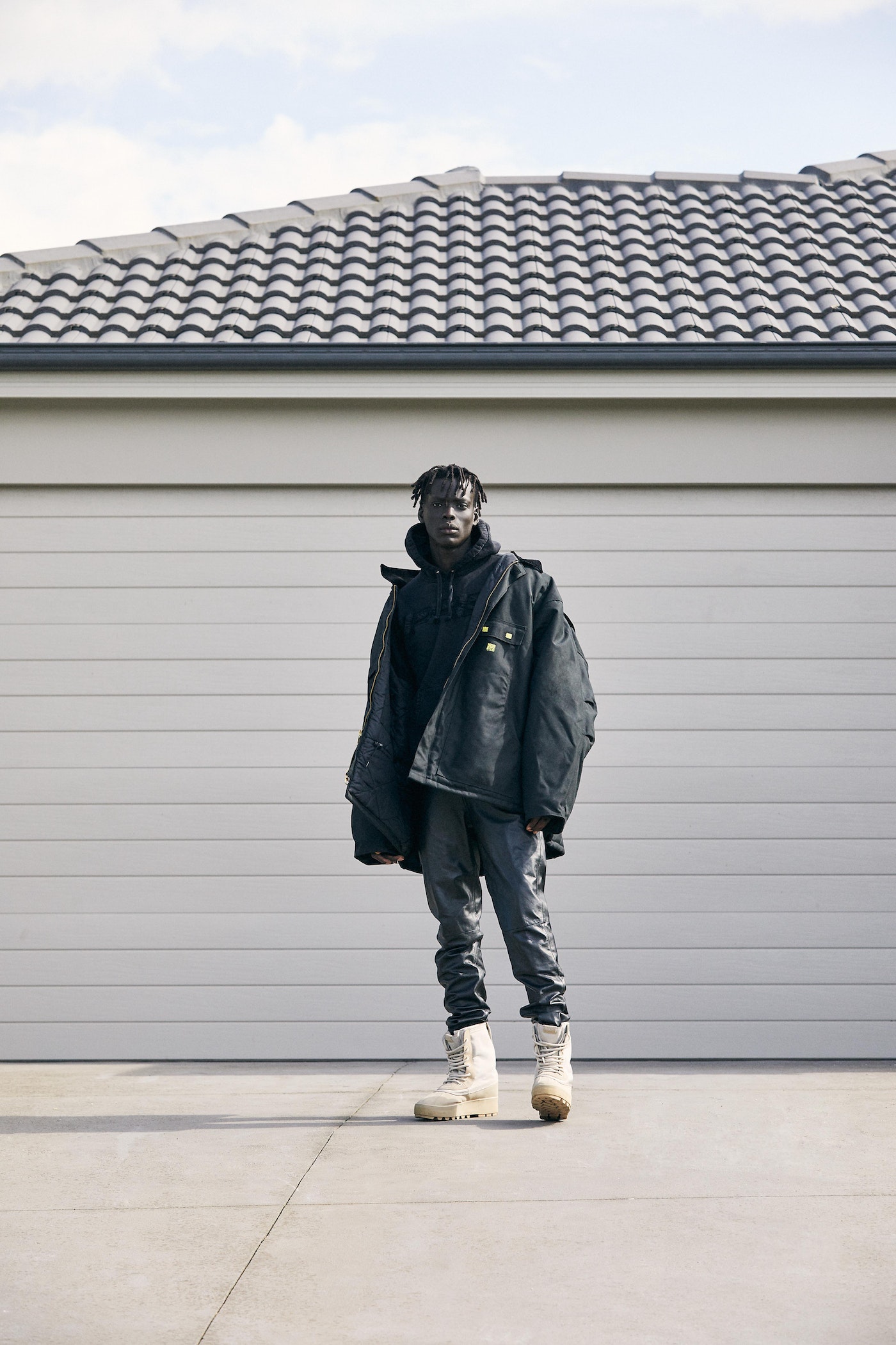 Full-length shot of a Black male wearing a black jacket, gray jeans, and cream-colored boots stands in front of a garage.