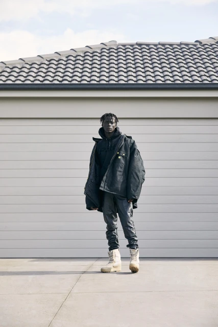 Full-length shot of a Black male wearing a black jacket, gray jeans, and cream-colored boots stands in front of a garage.