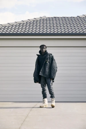 Full-length shot of a Black male wearing a black jacket, gray jeans, and cream-colored boots stands in front of a garage.