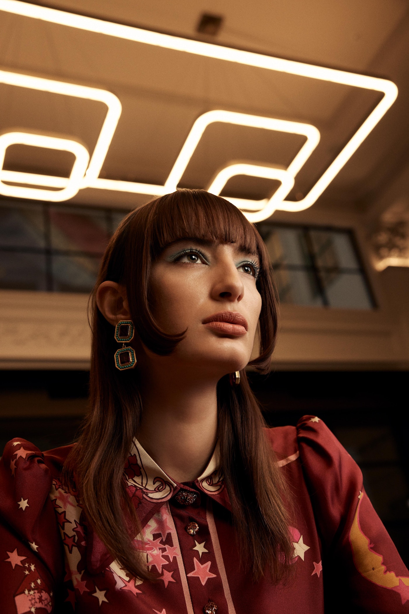 Eye-level portrait of a woman with side bangs, adorned in patterned clothing, gazing upwards with a neon light fixture above.