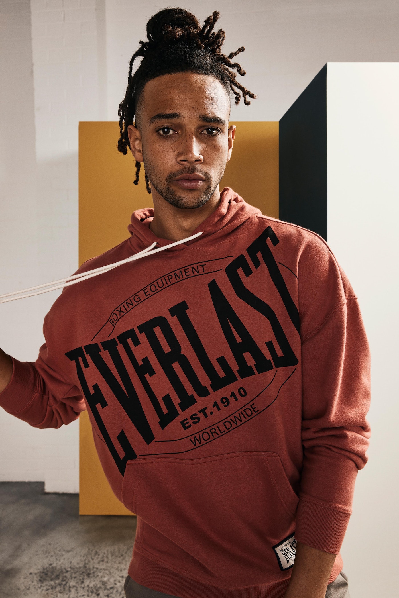 Man in a rust-colored Everlast hoodie in a casual studio setting with modern minimalist blocks in the background; indoor fashion shot.