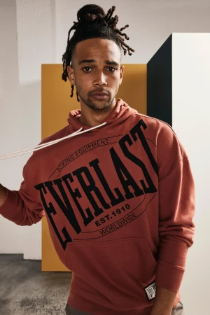 Man in a rust-colored Everlast hoodie in a casual studio setting with modern minimalist blocks in the background; indoor fashion shot.