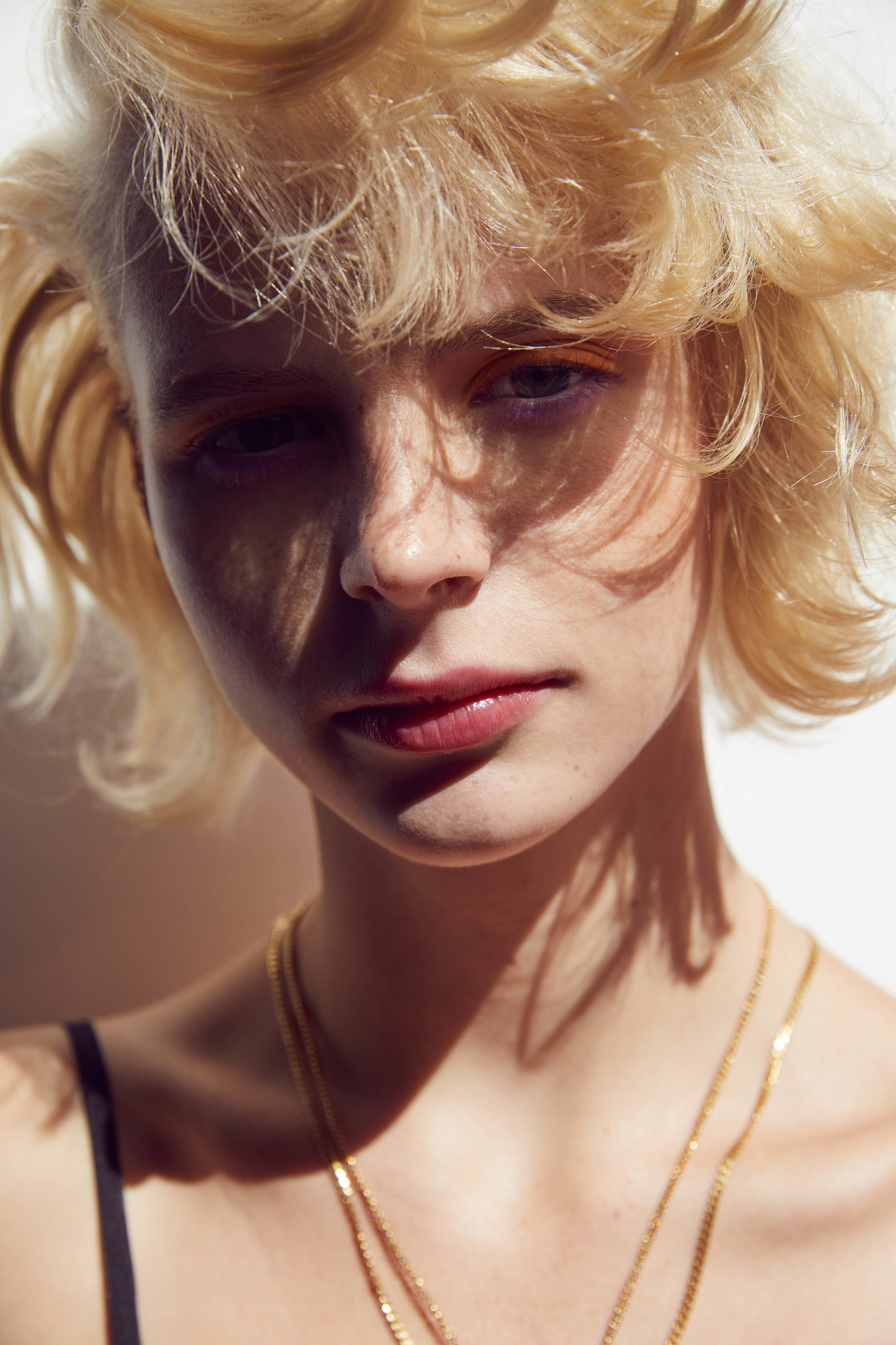 Close-up portrait of a woman with blonde hair, freckles, and gold chains against a plain background.