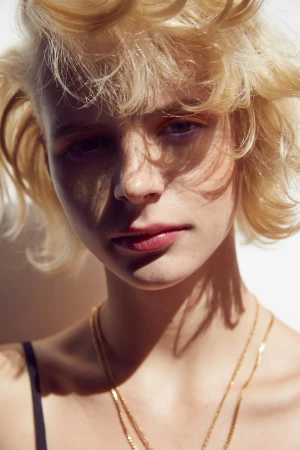 Close-up portrait of a woman with blonde hair, freckles, and gold chains against a plain background.