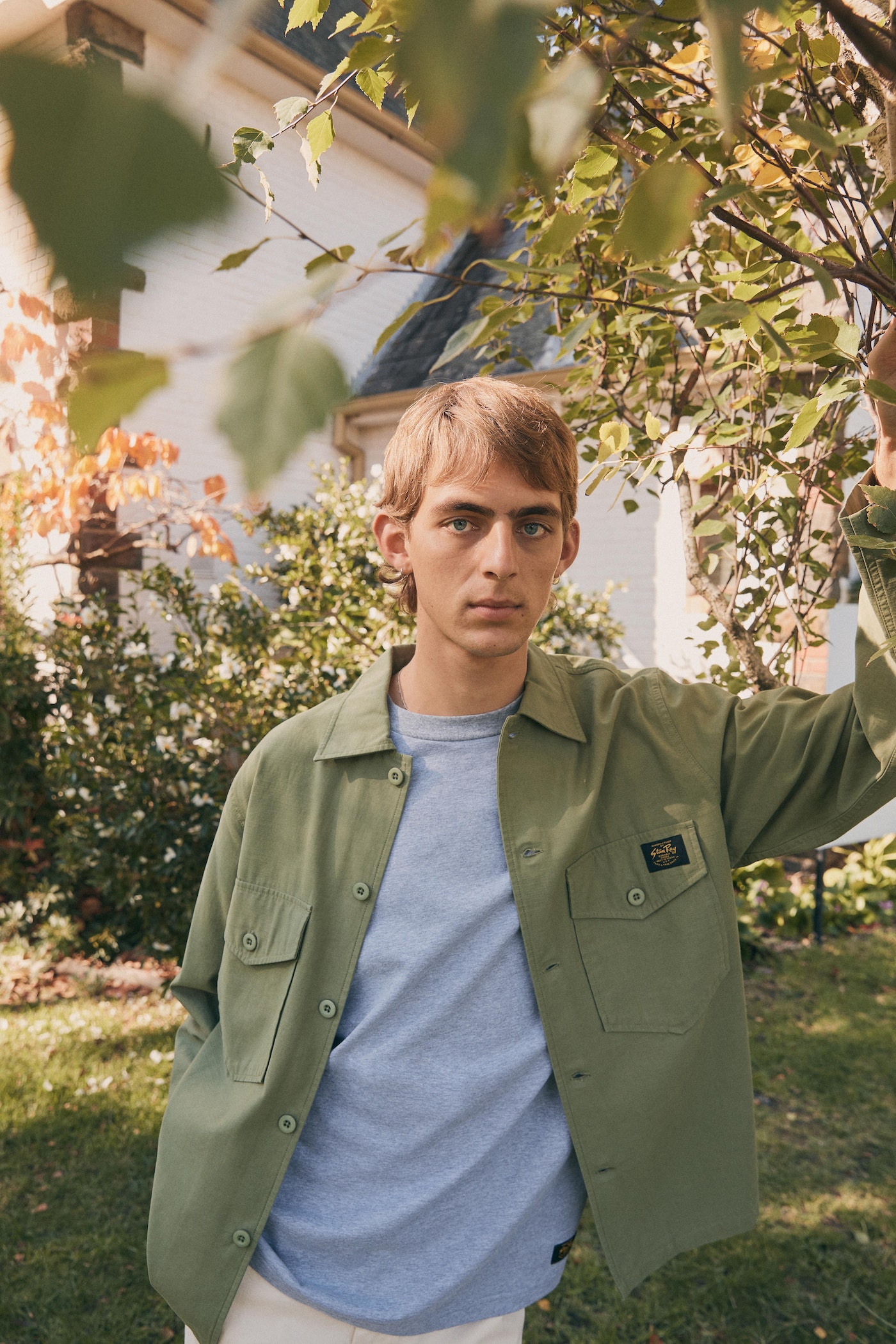 A fair-skinned young adult leans against a tree, wearing a sage-green jacket over a gray T-shirt, posing outside a house on a sunny day.