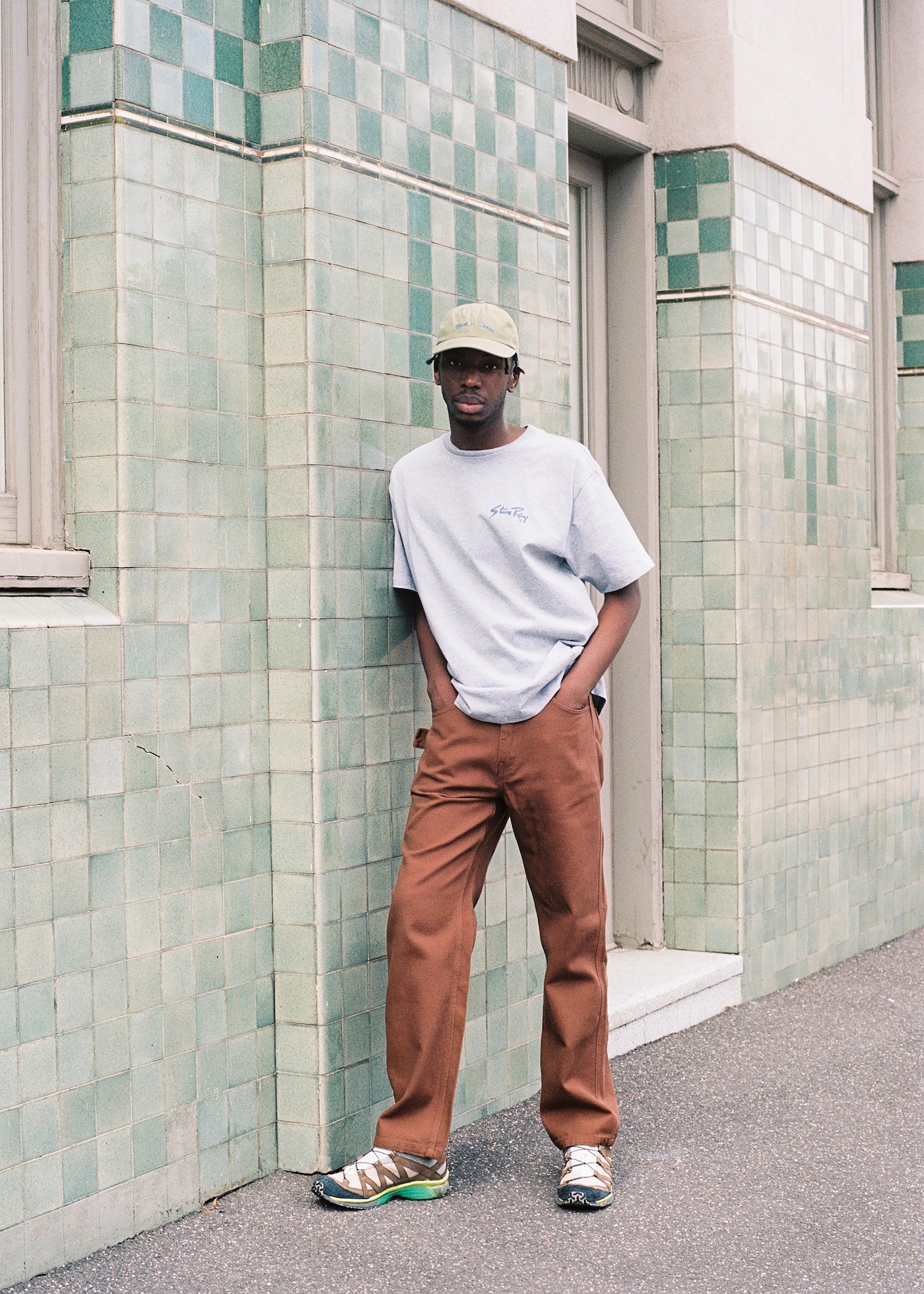 Full-length shot of a young Black male in casual attire, standing against a tiled building exterior, looking towards the camera.