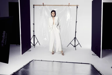 Model in a white pinstripe suit standing in a studio with light stands, black V-flats, and a transparent plastic sheet backdrop.
