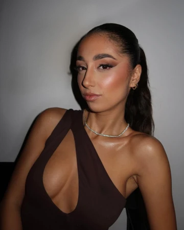 Close-up portrait of a woman wearing a dark brown one-shoulder cut-out top and a tennis necklace against a grey wall.