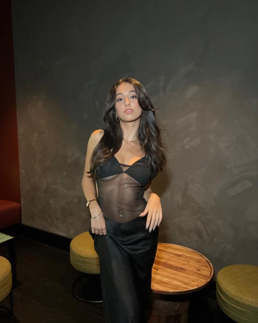 Portrait of a young woman with long brown hair in a black mesh dress. She is standing indoors with stools in the background.