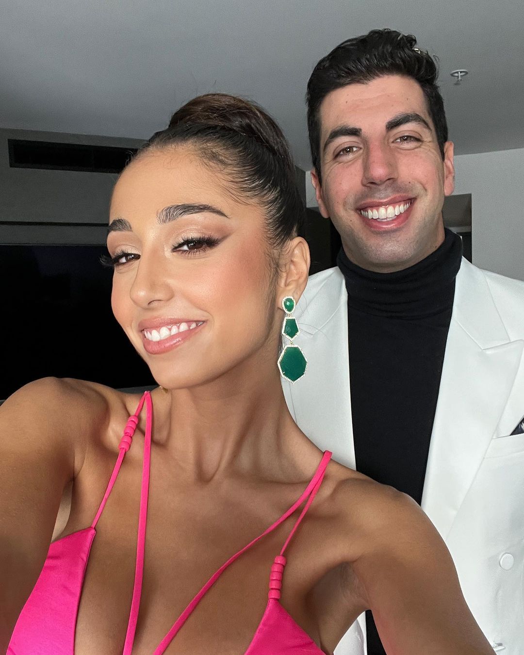 Smiling woman and man in bright fashionable attire pose in an indoor shot; she's in pink, he's in white and black.