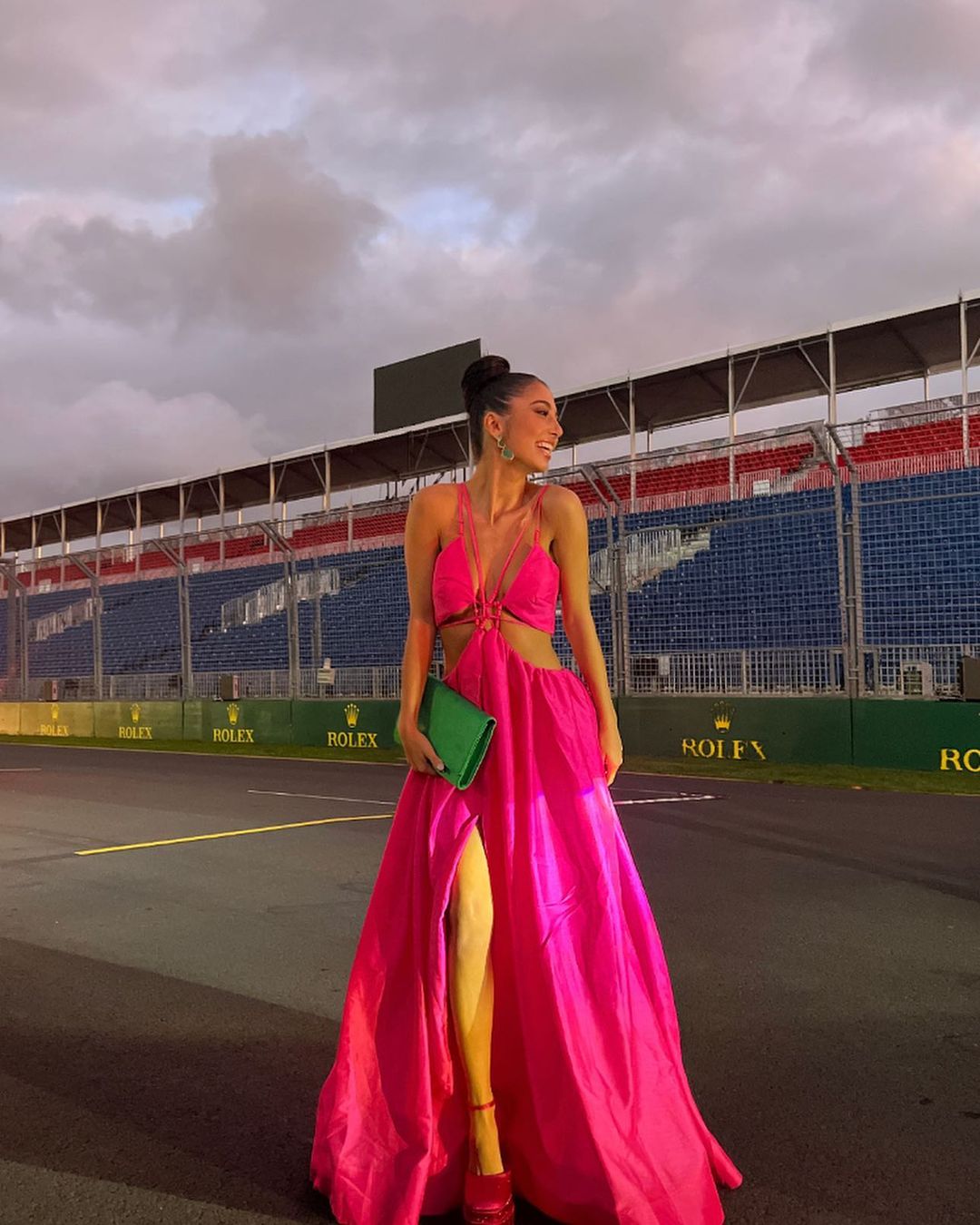 Woman in a pink dress holding a green clutch stands on a racetrack with empty stadium seating in the background on an overcast day.