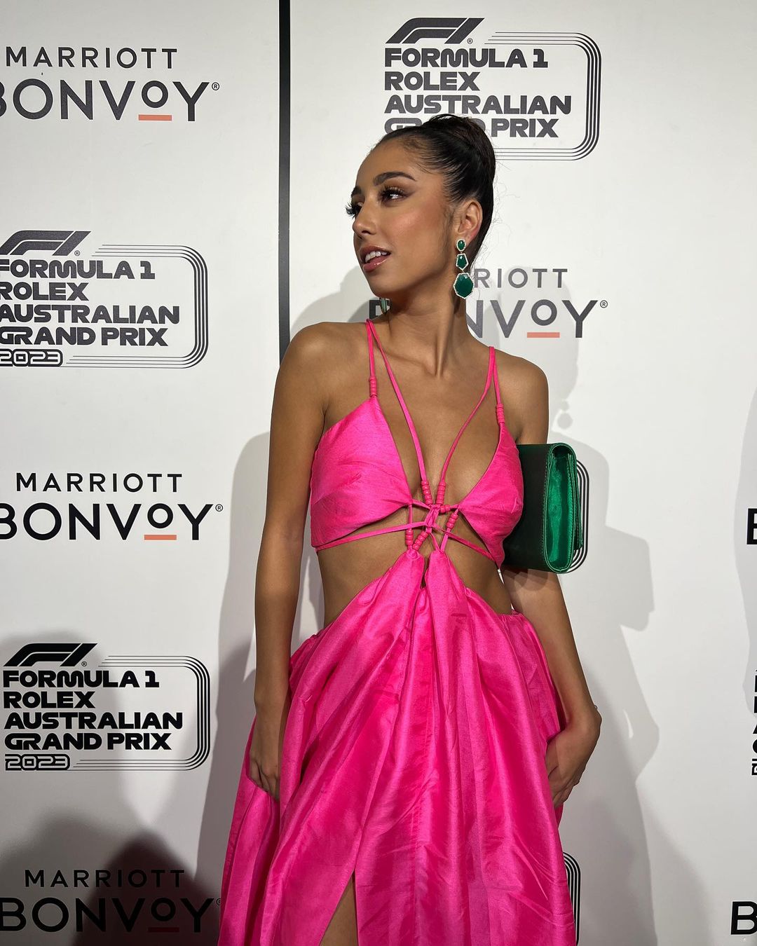 Woman in pink dress at Formula 1 Rolex Australian Grand Prix event; she holds a green clutch, and green jewel earrings are visible.