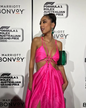 Woman in pink dress at Formula 1 Rolex Australian Grand Prix event; she holds a green clutch, and green jewel earrings are visible.