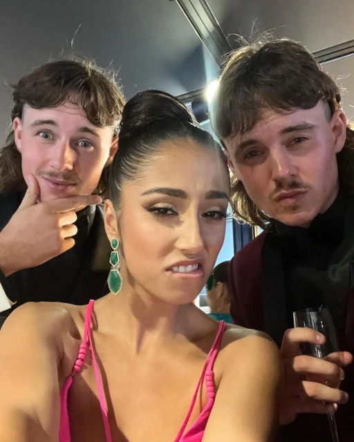 Three people pose indoors. Woman in pink dress with earrings in the center, two men with mullets, tuxedos with drinks on either side of her.