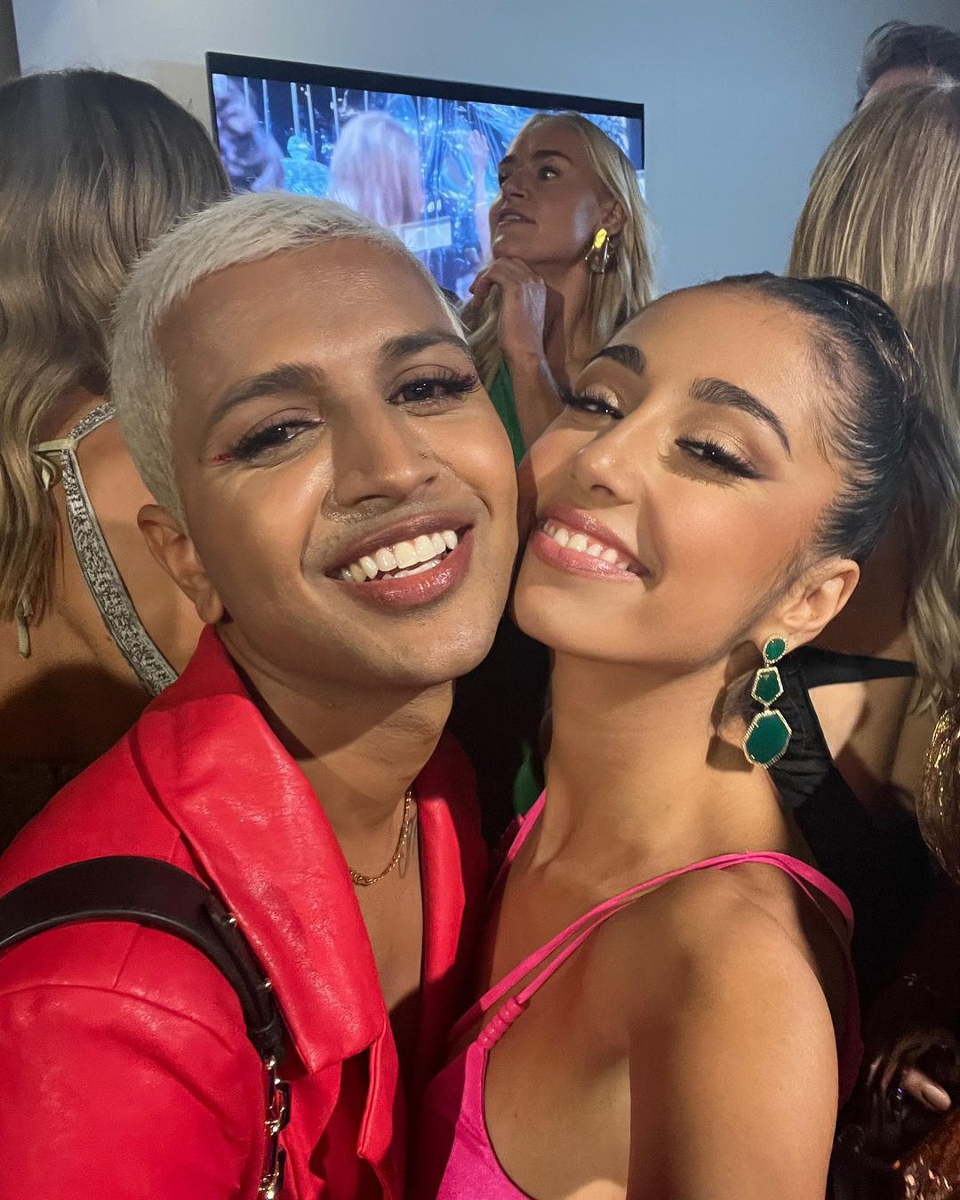 Two smiling adults pose indoors. One with short platinum hair wears a red jacket, the other with long hair wears a pink top and green earrings.