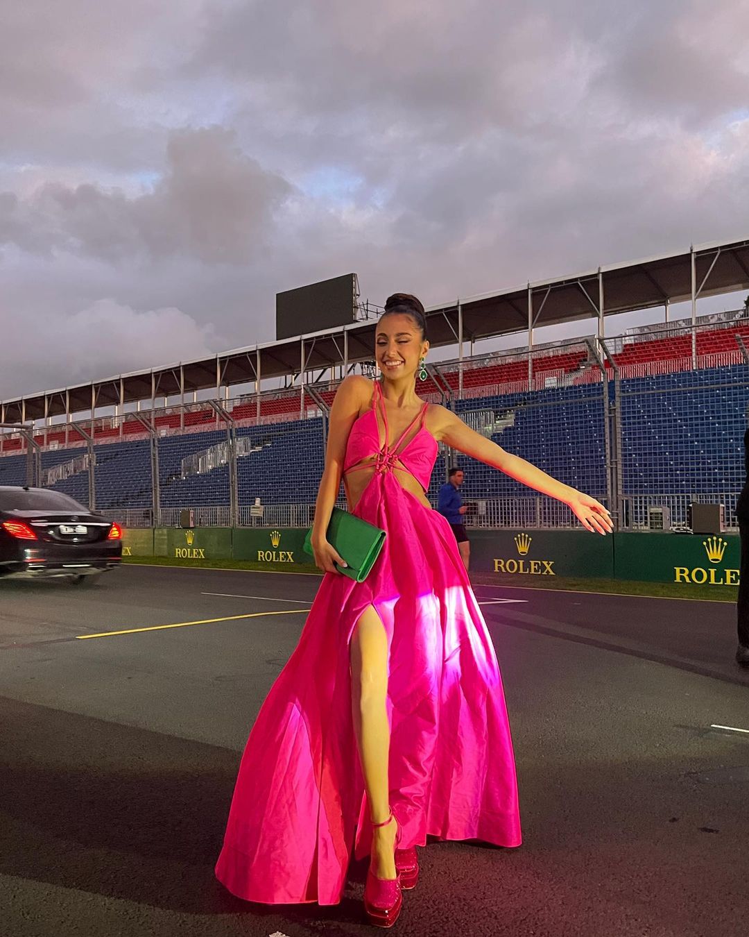 A woman in a bright pink gown and matching high heels poses at a racetrack. She has a green clutch and is smiling widely.