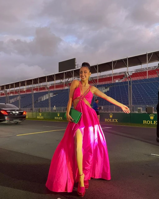 A woman in a bright pink gown and matching high heels poses at a racetrack. She has a green clutch and is smiling widely.