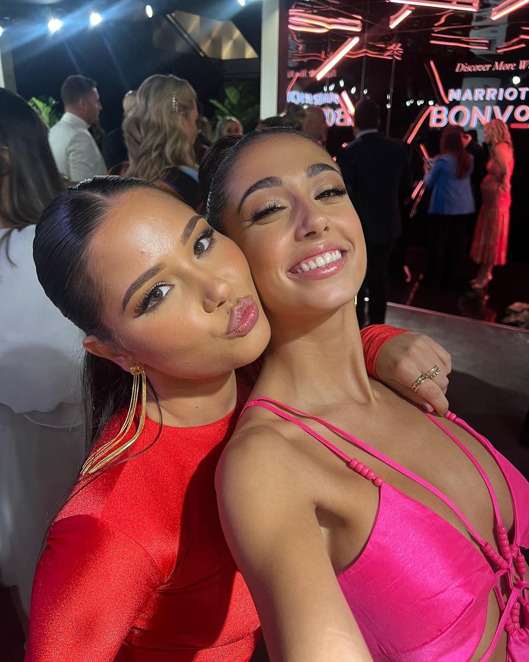 Two smiling women pose in front of a Marriott Bonvoy event backdrop. One wears a pink dress and the other a red dress.