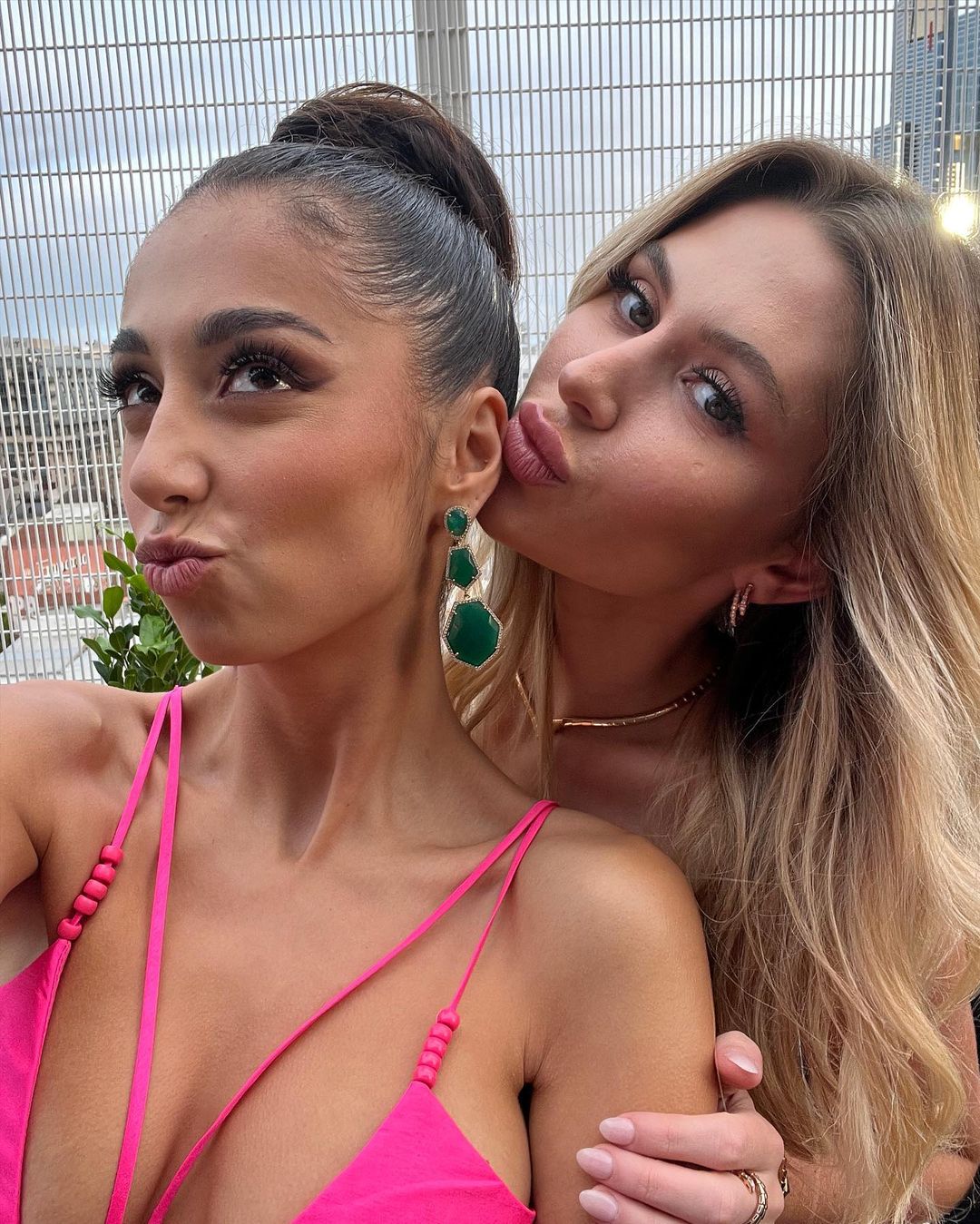 Two women pose closely outdoors. One kisses the other's cheek. The woman wears a pink top and green earrings against a city background.