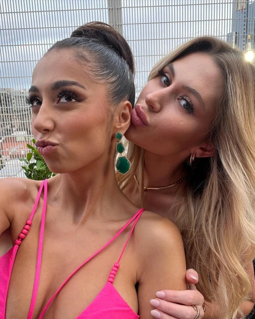 Two women pose closely outdoors. One kisses the other's cheek. The woman wears a pink top and green earrings against a city background.