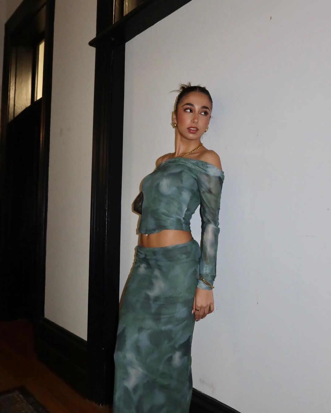 Woman in a green patterned off-the-shoulder long-sleeve top and matching skirt leaning against a white wall.