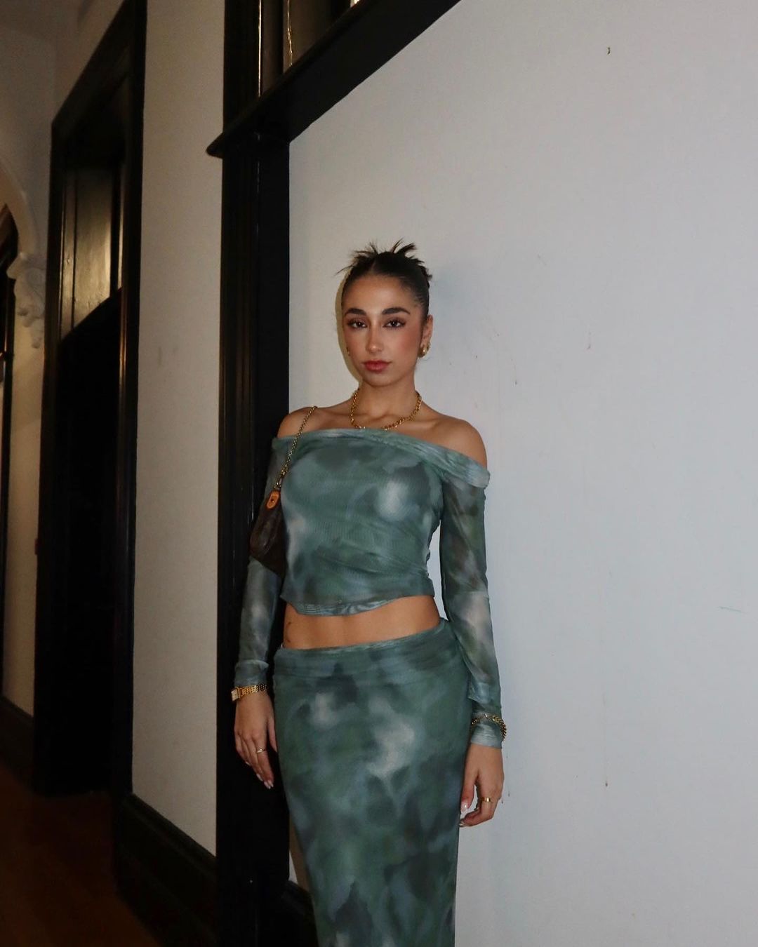 A woman poses indoors wearing a green patterned two-piece outfit with a handbag and jewelry for a fashion or lifestyle aesthetic.