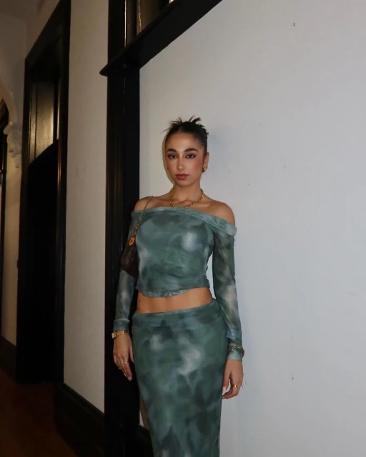A woman poses indoors wearing a green patterned two-piece outfit with a handbag and jewelry for a fashion or lifestyle aesthetic.