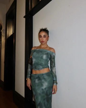 A woman poses indoors wearing a green patterned two-piece outfit with a handbag and jewelry for a fashion or lifestyle aesthetic.