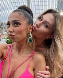 Two young women posing closely together, one with dark hair in an updo wearing a bright pink top and green earrings