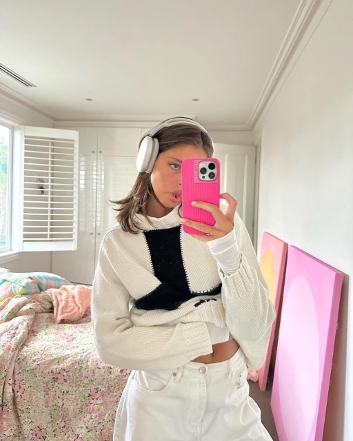 Young woman with headphones takes a mirror selfie indoors wearing a black and white sweater, white jeans, and holding a pink phone.