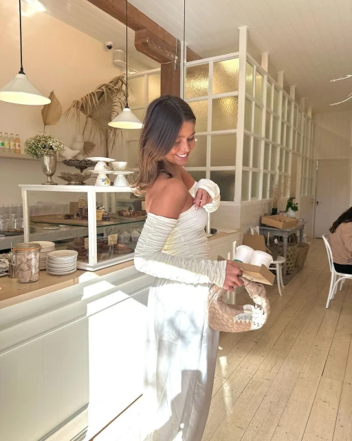 Indoor shot of a woman in a white dress smiling in a light-filled cafe, holding a handbag, with pastries visible on a display counter.