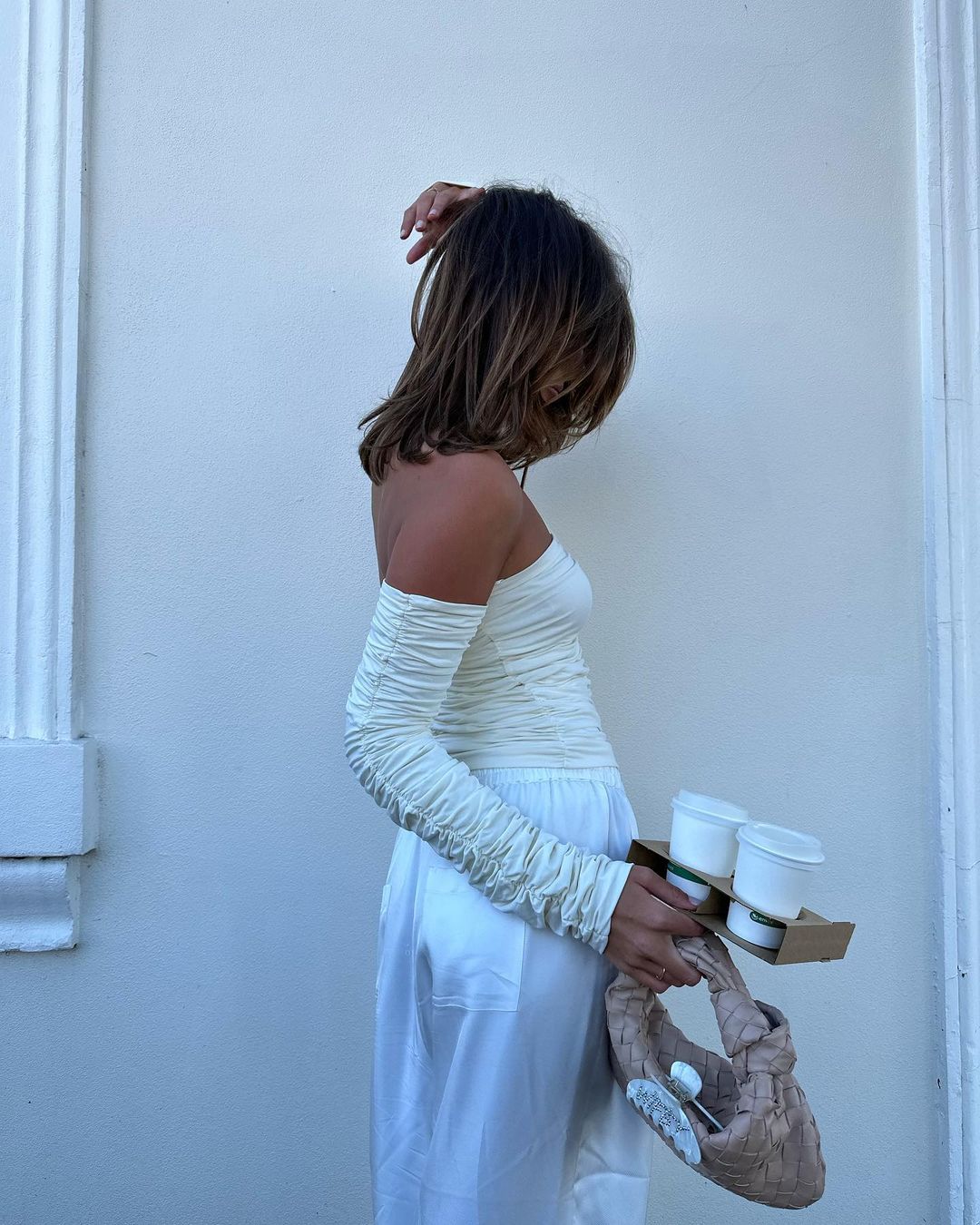 Adult woman in a white, off-the-shoulder top and matching wide-leg pants holds a drink carrier while posing near a light-colored wall.