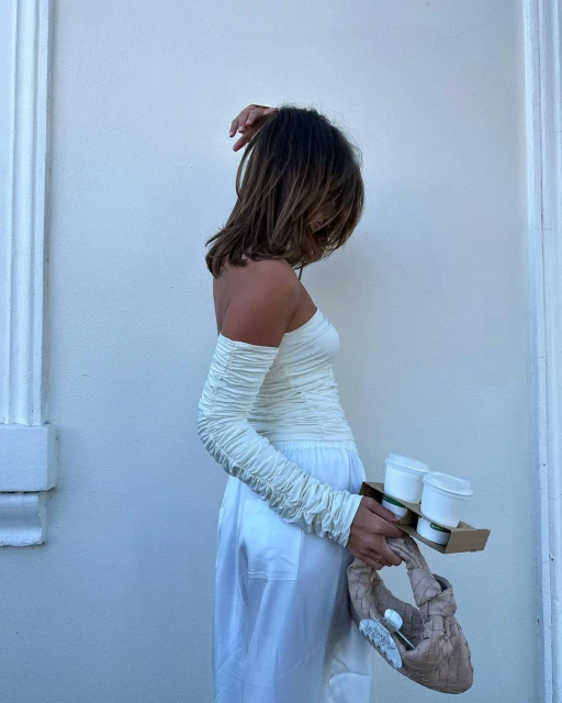 Adult woman in a white, off-the-shoulder top and matching wide-leg pants holds a drink carrier while posing near a light-colored wall.