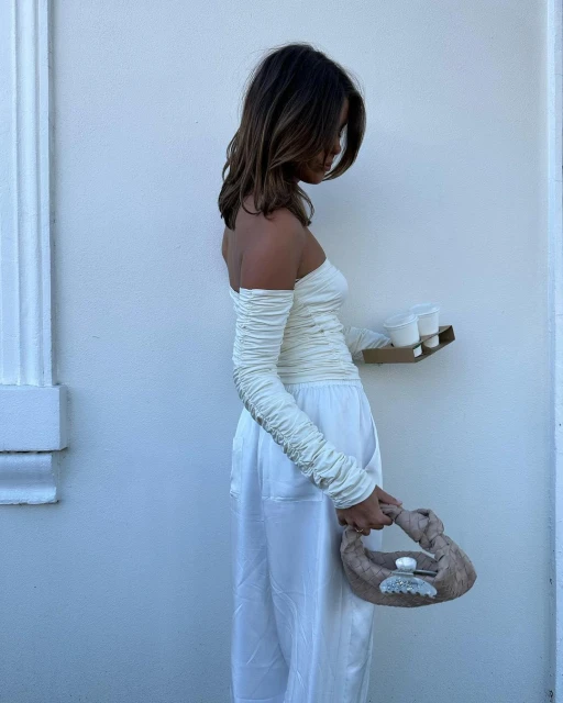 A fair-skinned young woman in a white outfit stands against a white wall, holding a brown handbag and a drink tray with two white cups.