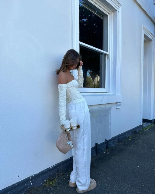 A woman in an off-shoulder top and white pants stands outside a white building, holding a drink carrier with three beverages.