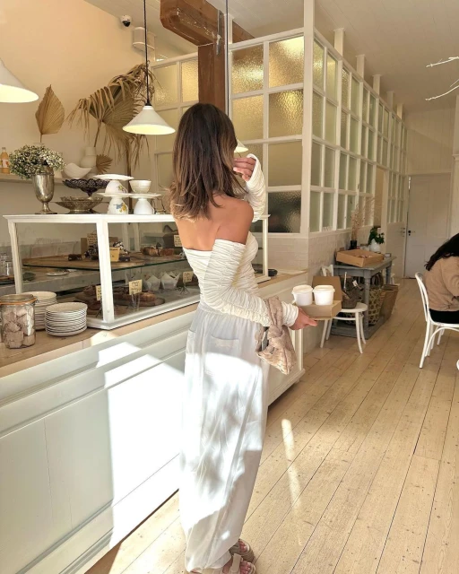 Back view of a woman in an off-the-shoulder top and white pants holding a tray with drinks in a cafe setting indoors.