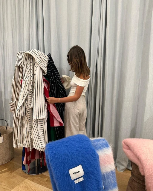 A woman browsing clothes on a rack, featuring striped shirts and soft blankets in blue and pink, with a neutral curtain backdrop.