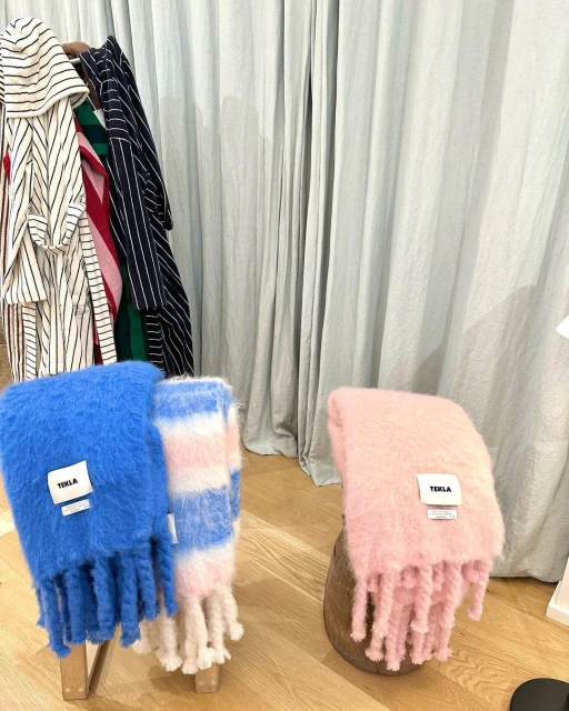 Three soft throws in blue, pink, and striped patterns are neatly displayed on light wooden stands against a soft grey curtain backdrop.