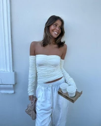 Smiling woman in white off-the-shoulder ruched top and matching sweatpants holding a coffee carrier against a white wall